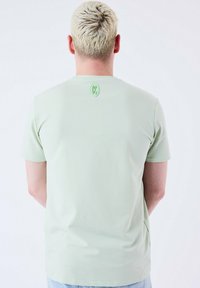 T-shirt verde chiaro a maniche corte, con texture liscia, caratterizzato da un piccolo logo in rilievo della stessa tonalità sul retro. Vestibilità casual.