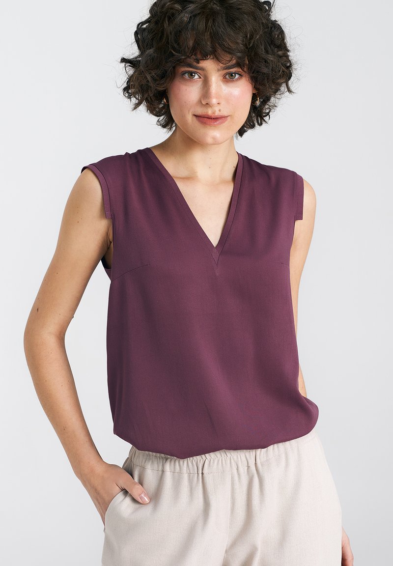 Top bez rękawów w kształcie litery V w głębokim burgundzie, wykonany z gładkiego materiału, z delikatnym detalem kieszeni. W połączeniu z jasnobeżowymi spodniami z elastycznego materiału.