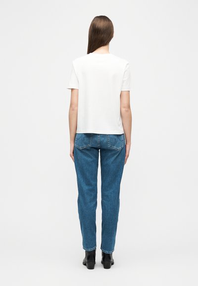 Γυναίκα με μακριά καστανά μαλλιά στέκεται με την πλάτη, φορώντας απλό λευκό κοντομάνικο μπλουζάκι, μπλε τζιν και μαύρα μποτάκια αστραγάλου.