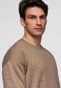 Beigen sävyinen neulepusero, jossa on kirjainkuvioinen tekstuuri, pyöreä pääntie ja kontrastinväriset sileät hihat. Pehmeä kangas, jossa hienovaraisia yksityiskohtia.