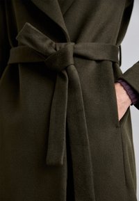 Cappotto di lana verde scuro con una texture morbida, dotato di una cintura annodata e di un colletto riflessato. Polsino di camicia viola visibile che spunta da sotto.