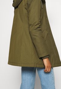 Olijfgroene gewatteerde jas met een capuchon, knoopsluiting aan de voorkant, zijzakken en ribgebreide manchetten. De stof lijkt lichtjes gestructureerd en duurzaam.