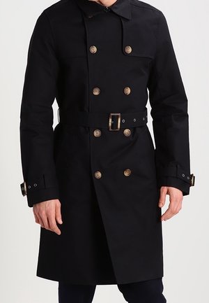 Mustan kaksirivisen trenssitakin, jossa ruskeat napit, vyötärölle solmittu vyö ja hihansuiden hihnat, pukeutunut henkilö yksinkertaista taustaa vasten.