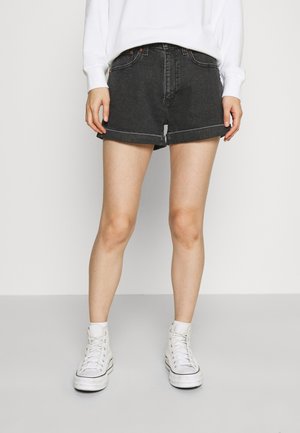 Pantalones cortos de mezclilla negra con puños doblados, que cuentan con un cierre de cremallera y botón, bolsillos laterales y un diseño ajustado, combinados con zapatillas altas blancas.