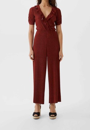 Combinaison rouge à manches courtes, col en V volanté, jambes larges et motif discret de petits points noirs. Portée avec des espadrilles à plateforme noires.