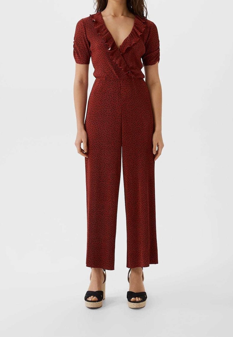 Combinaison rouge à manches courtes, col en V volanté, jambes larges et motif discret de petits points noirs. Portée avec des espadrilles à plateforme noires.