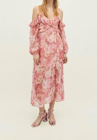 Vestido maxi floral en tonos de rosa con mangas con volantes sin hombros, material transparente y una silueta fluida. Combinado con tacones con tiras.