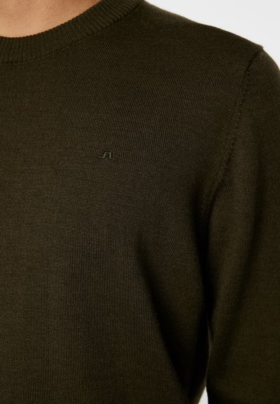 Mörkgrön stickad tröja med rund hals, med en subtil logotyp på bröstet. Tyget verkar slätt och något strukturerat.