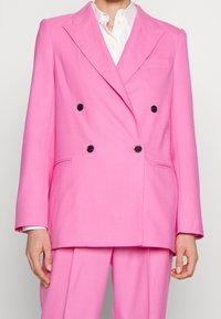 Blazer rosa doppiopetto con bottoni neri, rever a toppa e vestibilità tailored. Include una tasca sul petto e tasche laterali.