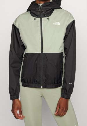 Person wearing a grey and black hooded zip-up jacket with elastic cuffs and logo on the chest, paired with light green leggings.