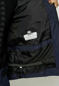 Mørkeblå jakke indvendig med sort foring, der har et ejendomsetiket, en lynlås-lomme og elastisk snor til justering af pasformen.