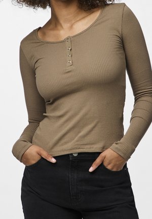 Femme portant un haut marron côtelé ajusté à manches longues avec trois boutons au niveau de l'encolure et un pantalon noir taille haute, les mains dans les poches.
