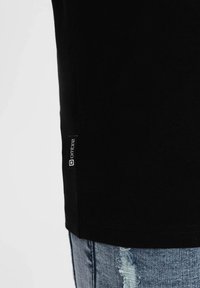 Schwarzes T-Shirt mit kurzen Ärmeln, das über eine glatte Stofftextur verfügt und ein kleines Markenlabel an der unter linken Naht angebracht ist.