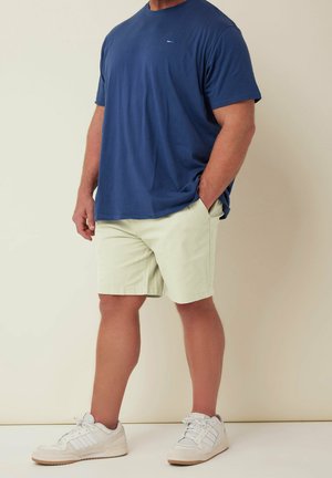 Uomo che indossa una maglietta blu navy, pantaloncini beige e scarpe da ginnastica bianche, in piedi con una mano in tasca su sfondo semplice.