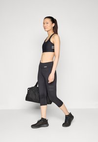 Must saada must spordivõimlemine rinnahoidja ja lühikesed legginsid kõrge vöökoht. Mudel hoiab musta jõusaali kotti ja kannab musti tekstuuriga spordijalatsid.