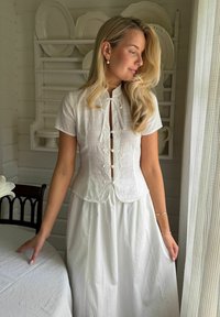 Femme blonde portant un chemisier blanc à manches courtes et une jupe se tient près d'une table avec du linge blanc, près d'une fenêtre avec des rideaux transparents.