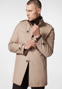 Beżowy płaszcz trencz z guzikami z przodu, naramiennikami i kieszeniami bocznymi. Gładki materiał, długość do kolan i wysoki kołnierz.