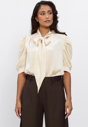 Woman wearing a cream satin blouse with a front bow and puffed sleeves, paired with high-waisted dark brown trousers, standing against white background.