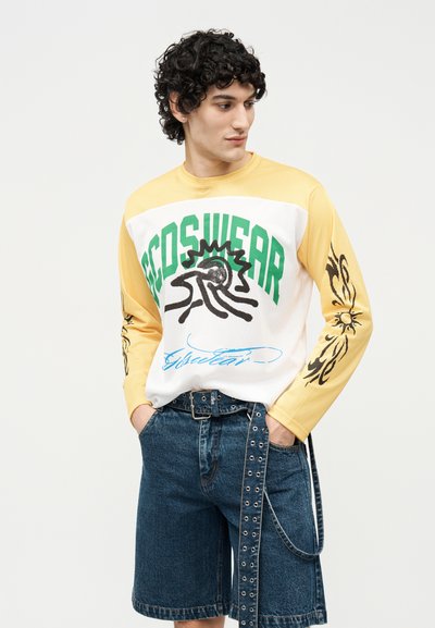 Νέος άνδρας που φοράει ένα κίτρινο και λευκό μακρυμάνικο μπλουζάκι με γραφικά και πράσινο κείμενο, με σκούρα μανίκια με floral σχέδιο, συνδυασμένο με μπλε τζιν σορτς και ζώνη με μεταλλικά κουμπώματα.