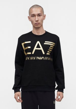 Homme au crâne rasé portant un sweat-shirt noir avec un grand logo doré EA7 Emporio Armani sur la poitrine, debout devant un fond blanc uni.