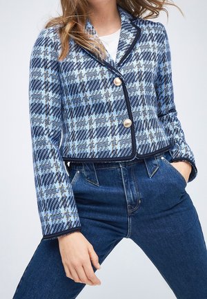 Vrouw draagt een blauw geruite blazer met gouden knopen over een wit topje en donkerblauwe high-waisted jeans met handen in de zakken.