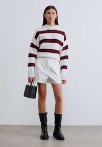 Ριγέ πουλόβερ πλεχτό σε λευκό με κόκκινες και μαύρες ρίγες, συνδυασμένο με λευκές σορτς και μαύρα μπότες μέχρι το γόνατο. Μικρή μαύρη τσάντα χειρός.