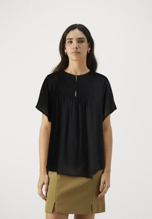 Blusa nera a maniche corte con design plissè, scollatura a forma di chiave e una superficie liscia. Abbinata a una gonna beige con spacchi laterali.