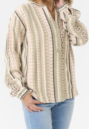 Vrouw draagt een blouse met lange mouwen met een patroon van verticale strepen in gedempte roze, groene en beige tinten, gecombineerd met lichtblauwe spijkerbroek.
