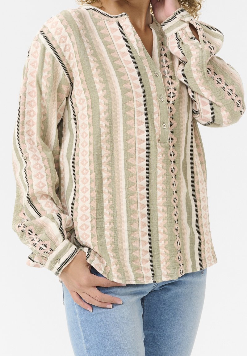 Femme portant un chemisier à manches longues à motifs avec des rayures verticales dans des tons rose pâle, vert et beige, associé à un jean bleu clair.
