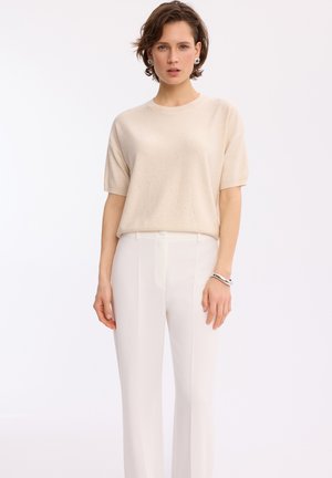 Kobieta o krótkich brązowych włosach, ubrana w beżowy sweter z krótkim rękawem, białe spodnie z wysokim stanem, srebrne kolczyki i srebrną bransoletkę, stojąca na białym tle.