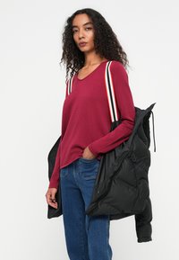 Bordeauxkleurig longsleeve shirt met een V-hals, gecombineerd met een blauwe spijkerbroek en een zwarte pufferjack dat over de schouder wordt gehouden. Gestreepte bandjes in rood en wit.