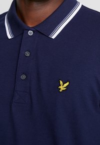 Marinblå polo skjorta med vitrandig krage, treknappars placket och en gul broderad fågel-logotyp på bröstet. Strukturerad tyg.