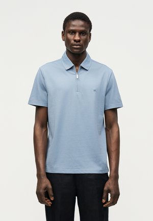 Mann trägt hellblaues kurzärmeliges Polo mit Reißverschluss und schwarze Hose, steht vor einfachem hellem Hintergrund.