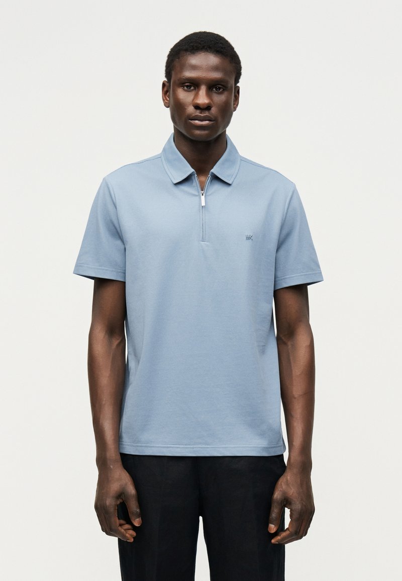 Άνδρας που φοράει ανοιχτό μπλε πόλο με κοντά μανίκια και φερμουάρ και μαύρο παντελόνι, στέκεται μπροστά σε απλό φωτεινό φόντο.