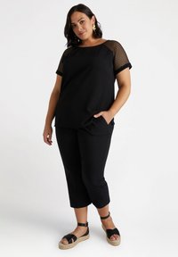 Haut noir avec des manches en maille, associé à un pantalon noir cropped. La tenue présente une coupe décontractée ; des sandales avec une semelle tissée ajoutent un détail.