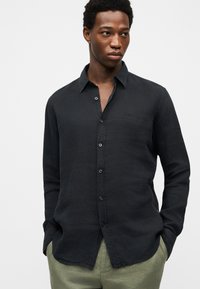 Νεαρός άνδρας που φοράει μαύρο πουκάμισο με κουμπιά και μακριά μανίκια και ανοιχτοπράσινο παντελόνι, με τα χέρια στις τσέπες, κοιτάζοντας ελαφρώς προς τα πλάγια.