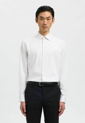 Man die recht vooruit kijkt, gekleed in een witte blouse met knopen, ingestopt in een zwarte broek met een zwarte riem.