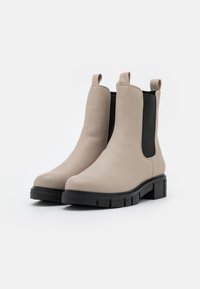 Bottines Chelsea en cuir beige avec des panneaux élastiques latéraux noirs et des languettes de tirage, présentant une semelle en caoutchouc noir épaisse avec des détails texturés.