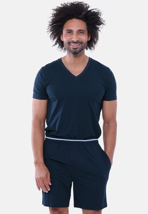 Man met krullend haar glimlachen, draagt een donkerblauw V-hals T-shirt en bijpassende korte broek met handen in de zakken tegen een effen achtergrond.