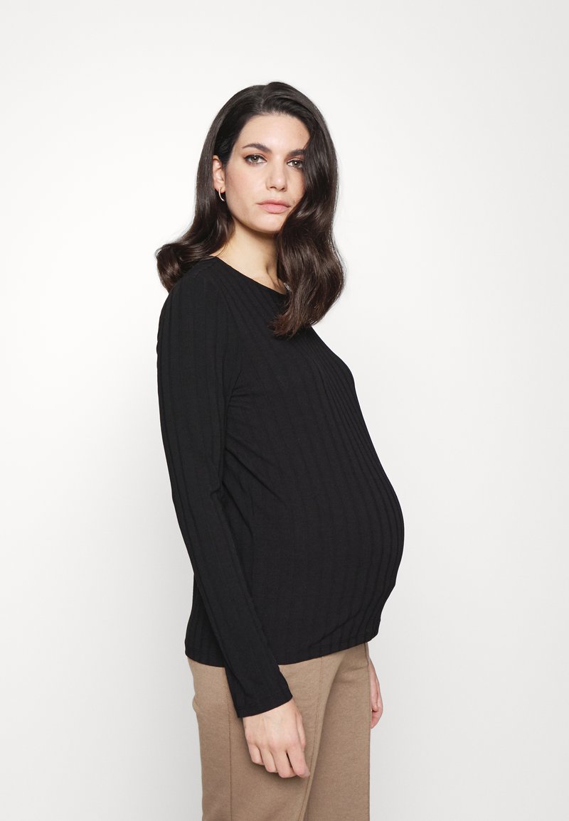 Camisola preta de mangas longas com decote redondo, corpo justo e uma silhueta visivelmente grávida, combinada com calças bege.