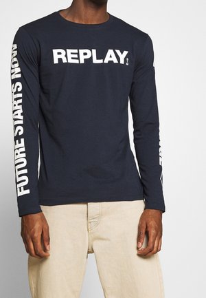 Homme portant une chemise à manches longues bleu marine avec "REPLAY" sur la poitrine et "FUTURE STARTS NOW" sur la manche droite, associée à un pantalon beige.