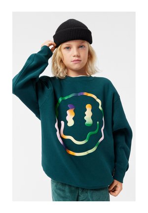 Felpa verde con una grafica di un sorriso colorato. Presenta una vestibilità comoda e polsini a coste. Indossata con un berretto di maglia nero.