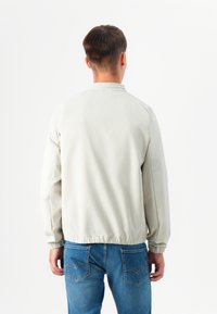 En ung mann står med ryggen til, iført en lys grå jakke og blå jeans, mot en enkel hvit bakgrunn.