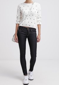 Suéter blanco de punto con lunares negros, combinado con jeans ajustados negros y zapatillas blancas. El modelo lleva un pequeño bolso cruzado.