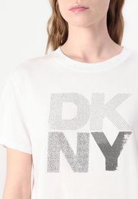 Balta marškinėlių su dideliu "DKNY" logotipu sidabriniais akmenukais, su apvaliu iškirpimu ir trumpomis rankovėmis. Lygus medvilnės audinys.