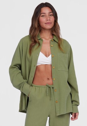 Olijfgroene overhemd met knopen, gemaakt van een geweven stof, met een vo zak en houten knopen, gedragen over een witte crop top en bijpassende broek.