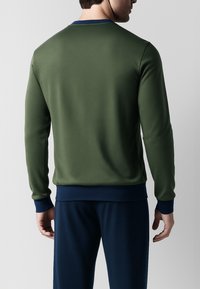 Felpa verde oliva con scollo rotondo, polsini e orlo blu scuro a contrasto, e tessuto liscio e leggero. Vista posteriore che mostra una forma aderente.