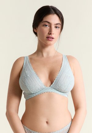 Frau mit dunklen Haaren, die ein hellblaues Spitz-Bralette und passende Unterwäsche vor einem schlichten beigen Hintergrund trägt.