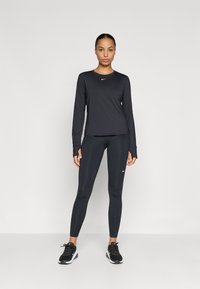 Μαύρο αθλητικό μπλουζάκι με μακριά μανίκια και κολάν, και τα δύο κατασκευασμένα από απαλό ύφασμα. Διαθέτει λευκές λεπτομέρειες με λογότυπα. Το μοντέλο φορά μαύρα αθλητικά παπούτσια.