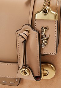Dettaglio di una borsa in pelle beige con accessori dorati, un'etichetta decorativa con un design di cavallo e carrozza e accenti di cucitura in contrasto.